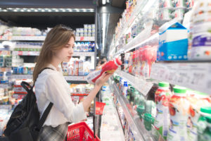 Giovane donna esamina l'etichetta di uno yogurt da bere, kefir o latte davanti al banco frigo di un supermercato