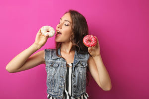 Giovane donna tiene in mano due ciambelle donut colorate mentre sta per addentarne una