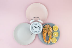 Due piatti vuoti e uno con salmone e fette di limone, con al centro una sveglia; concept: digiuno intermittente
