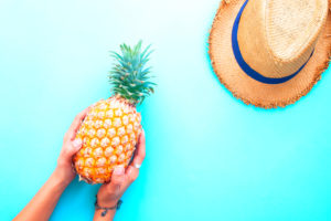 Mani femminili tengono ananas su sfondo azzurro, accanto a cappello di paglia; concept: vacanze, estate, frutta esotica
