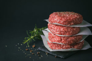 Quattro hamburger di bovino crudi impilati e divisi da fogli di carta forno, circondati da sale, pepe e rosmarino