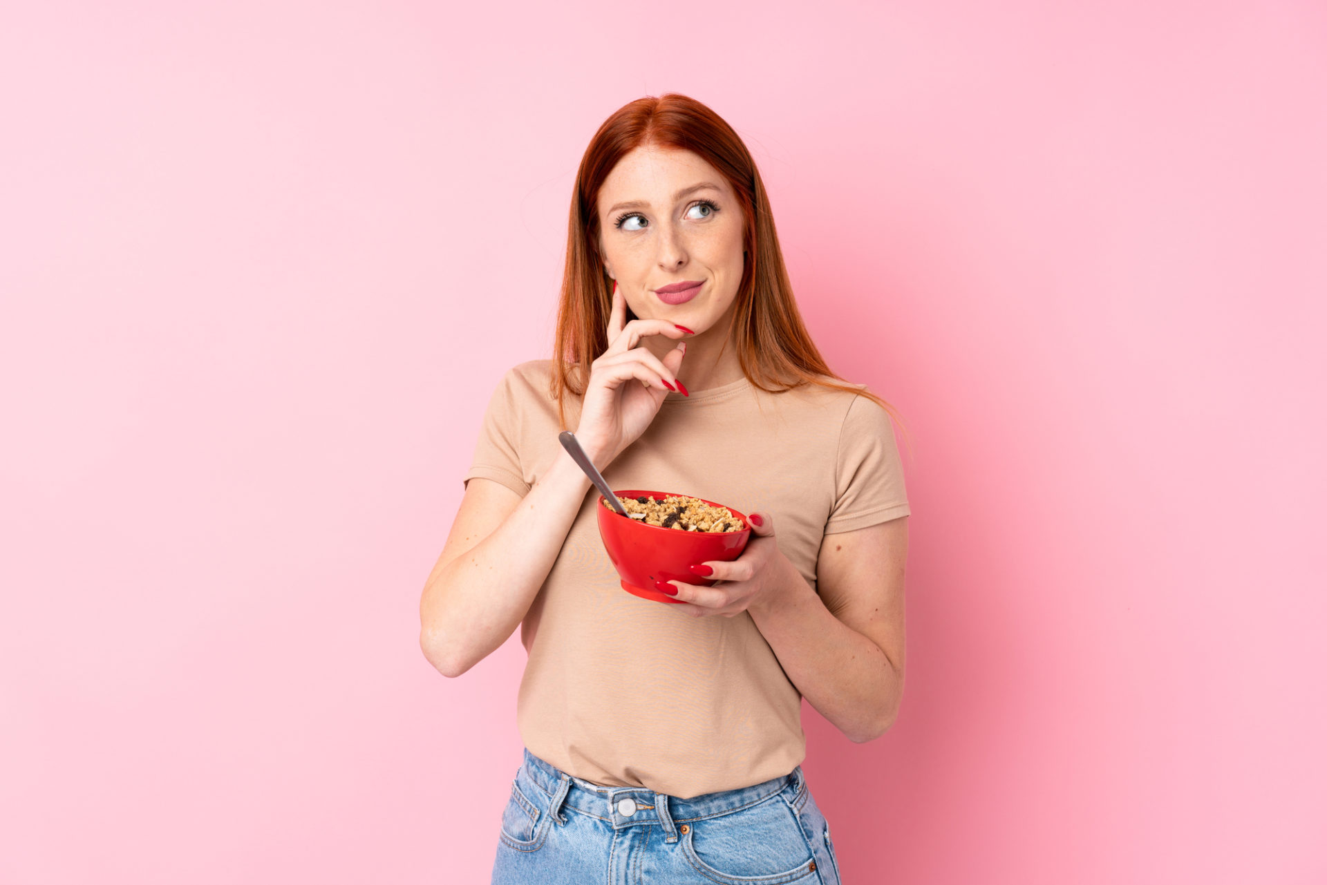 Giovane donna dai capelli rossi su sfondo rosa isolato che tiene in mano una ciotola di cereali e pensa
