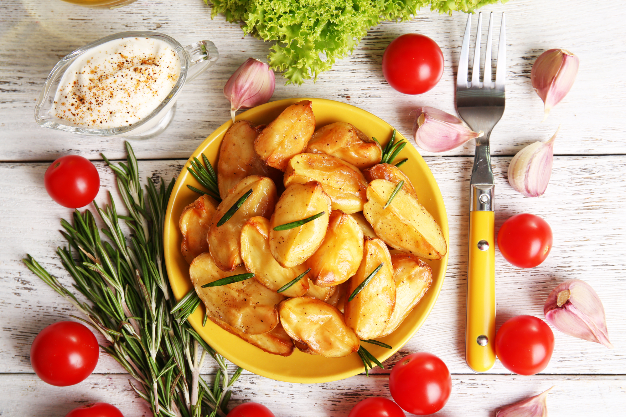 Piatto di patate a spicchi con rosmarino circondato da, da sinistra a destra: rami di rosmarino, ciotola di salsa, spicchi d'aglio, insalata, pomodorini e forchetta