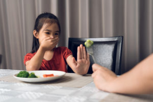 Bambina si rifiuta di mangiare broccolo proposto da un genitore, seduta a tavola davanti a un piatto di verdure; concept: neofobia alimentare