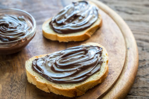 Due fette di pane con crema spalmabile al cacao e nocciole accanto a ciotola con crema su un piatto di legno; concept: Nutella