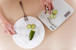 Una mano tiene un cetriolo, l'altra un coltello sullo sfondo di un piatto bianco. dieta pesare disturbi del comportamento alimentare anoressia bulimia vigoressia ortoressia