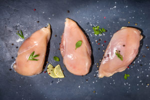 Tre petti di pollo crudi con sale, pepe, rosmarino e limone su sfondo scuro; concept: carne cruda