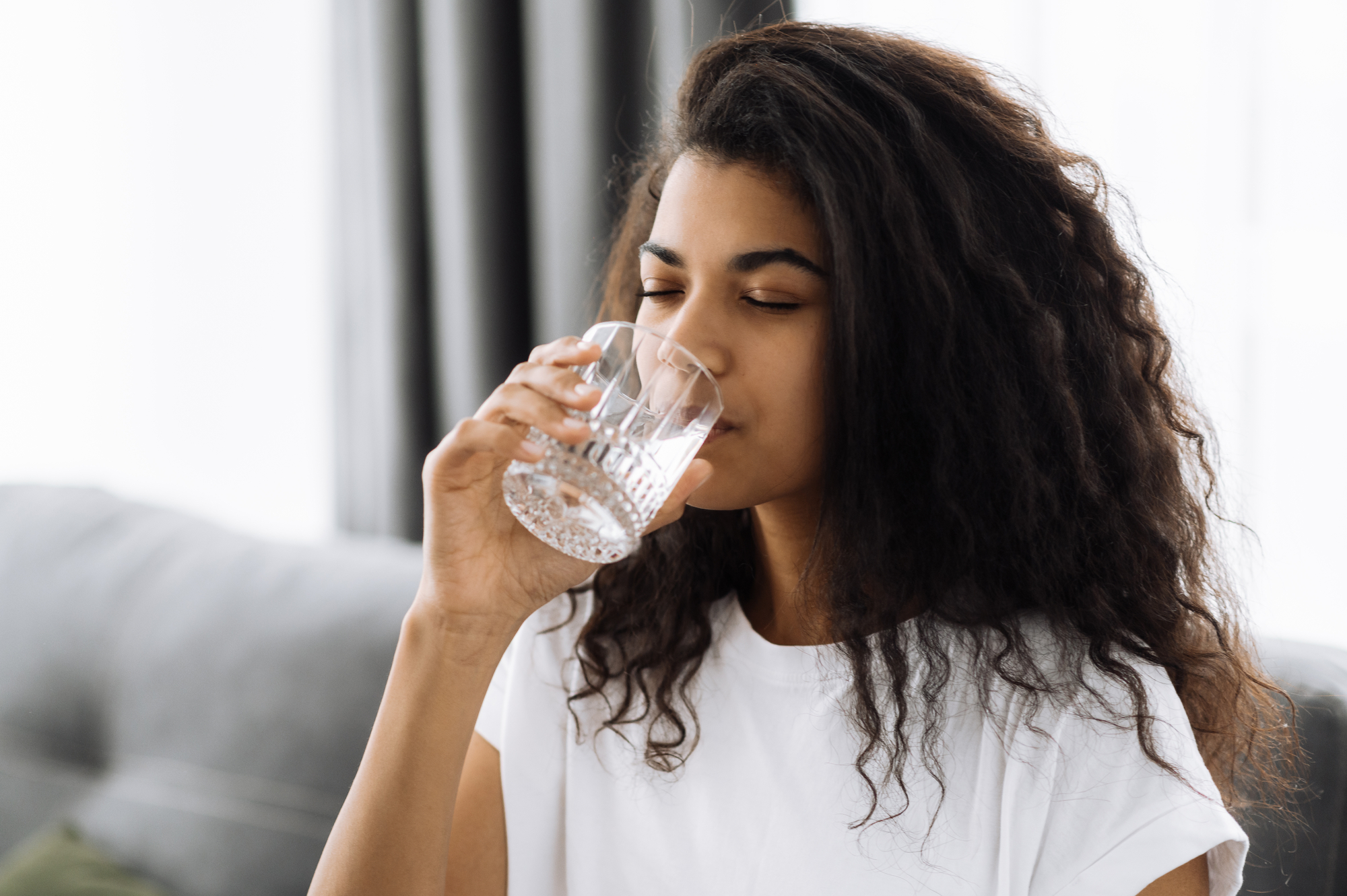 Stile di vita sano. Giovane donna afroamericana beve un bicchiere d'acqua mentre è seduta sul divano di casa. Bella donna sana segue uno stile di vita sano idratazione 