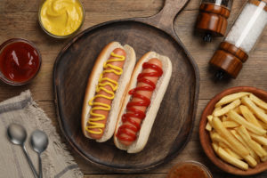 Due hot dog con senape e ketchup su un piatto di legno con patatine fritte e salse; concept ultra processati, junk food