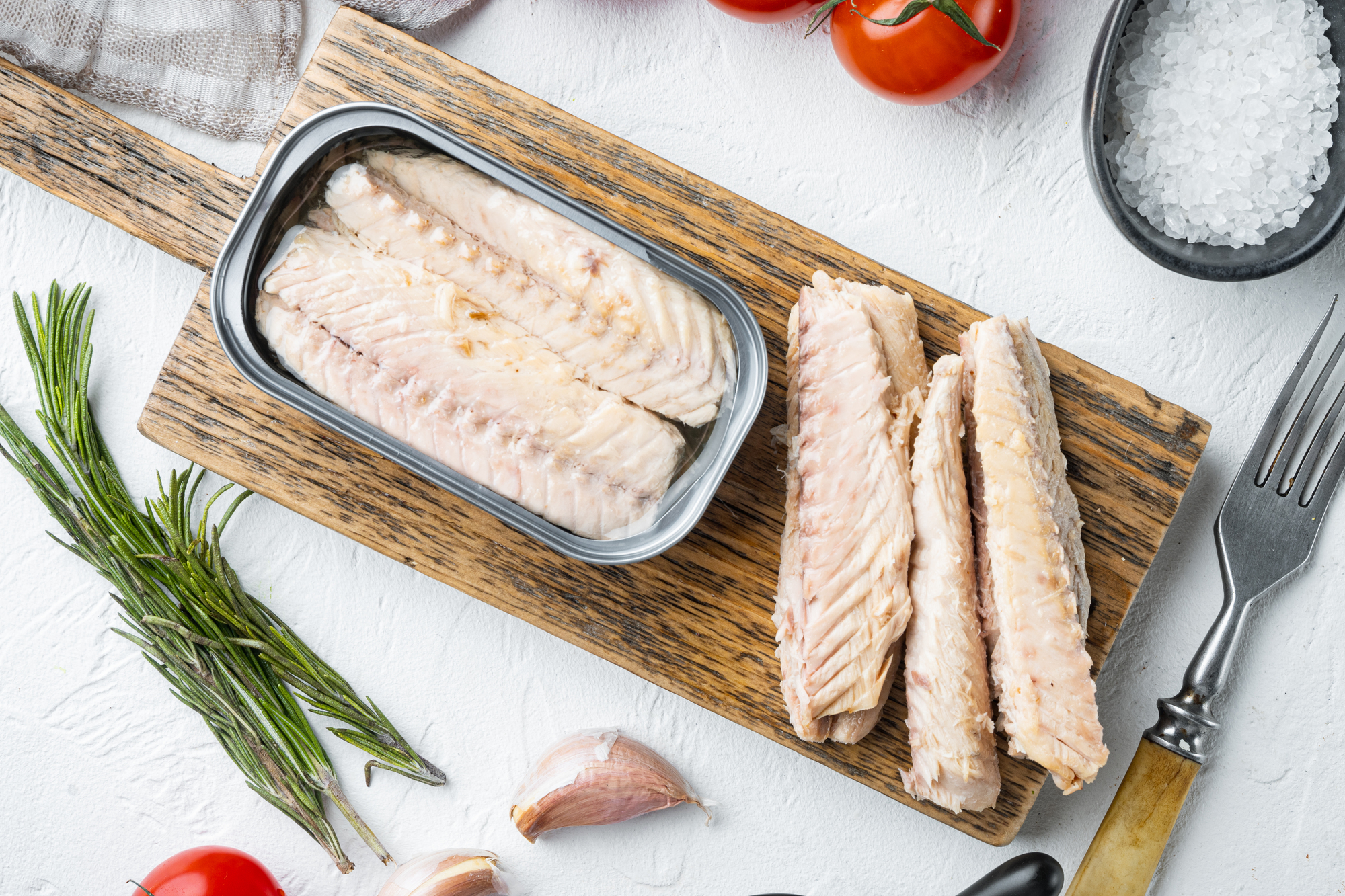 filetti di sgombri in scatola su tagliere di legno, circondati da erbe aromatiche, aglio, pomodorini, e sale; concept: pesce in scatola