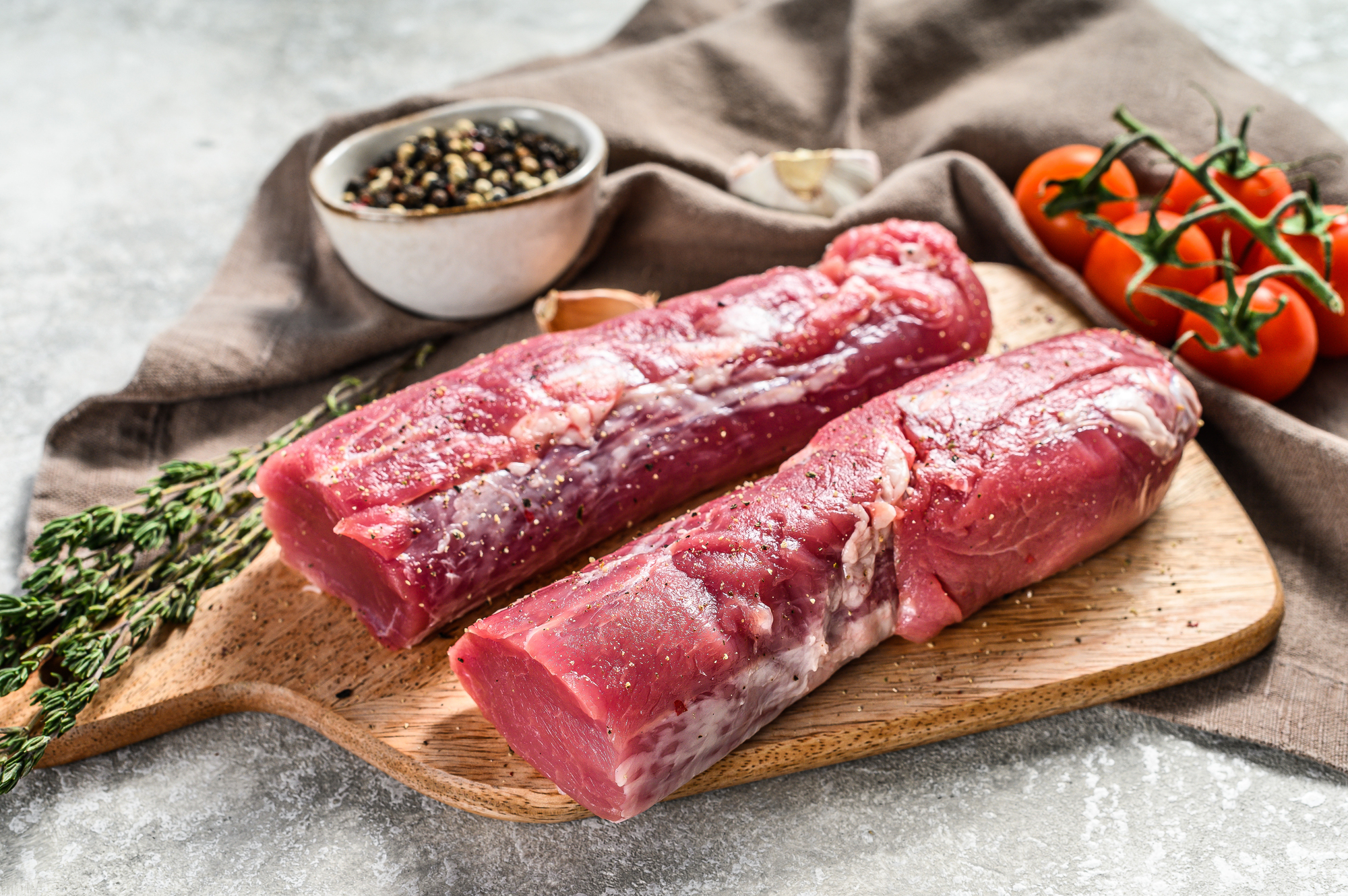 Filetto di suino su tagliere di legno accanto a erbe aromatiche, pomodorini e ciotola di pepe