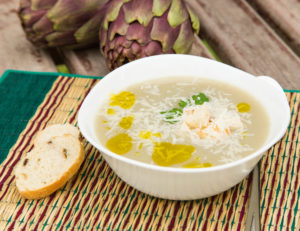 Piatto di zuppa di carciofi con formaggio grattugiato e, olio e crostini di pane; carciofi sullo sfondo