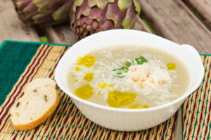 Piatto di zuppa di carciofi con formaggio grattugiato e, olio e crostini di pane; carciofi sullo sfondo