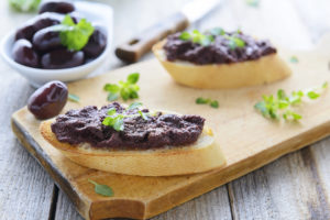 Crostini di pane con paté di olive su un tagliere di legno con erbe e ciotola di olive nere sullo sfondo