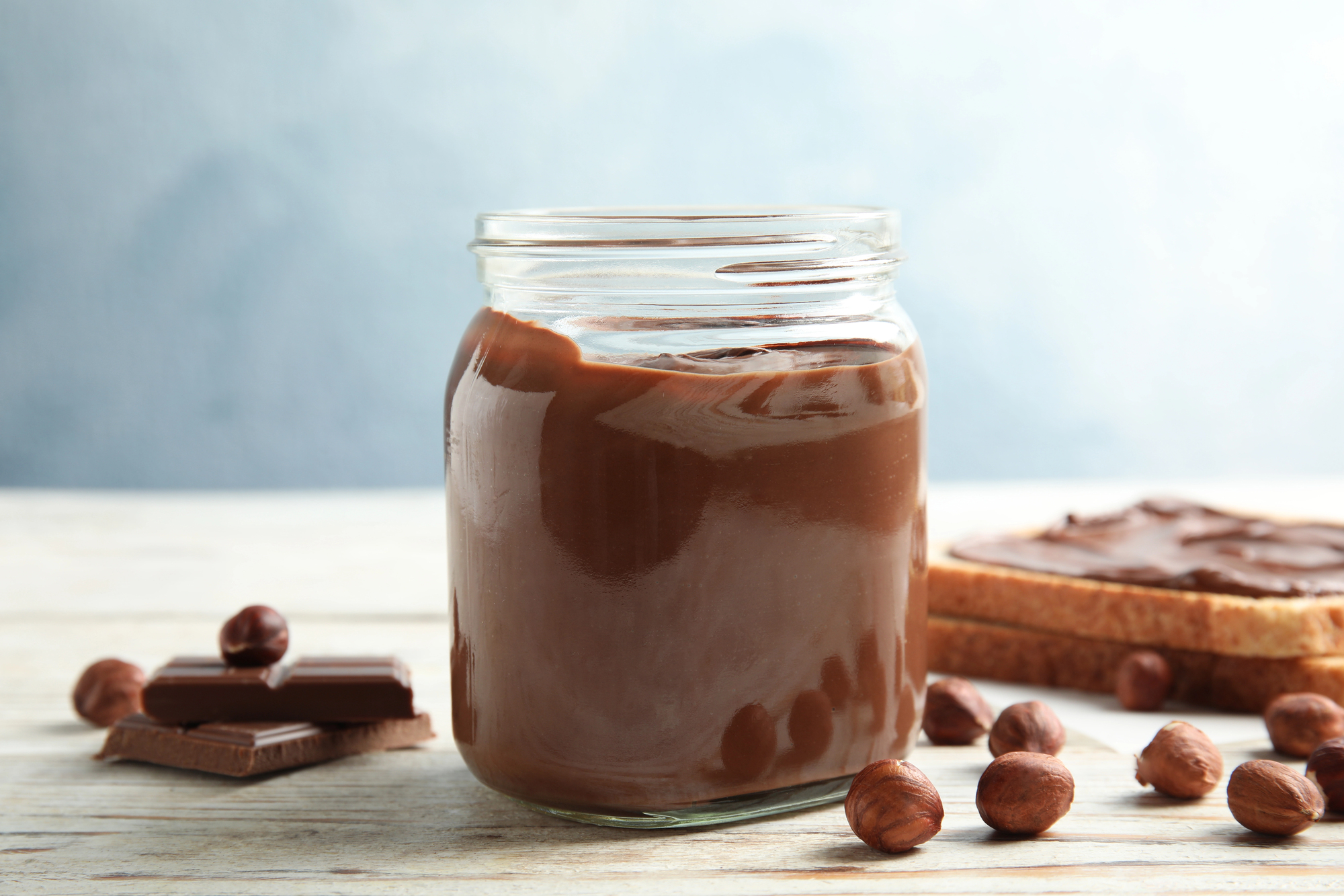 Un vasetto di crema spalmabile al cioccolato e nocciola tipo Nutella accanto a fette di pane con crema, nocciole intere e quadretti di cioccolato