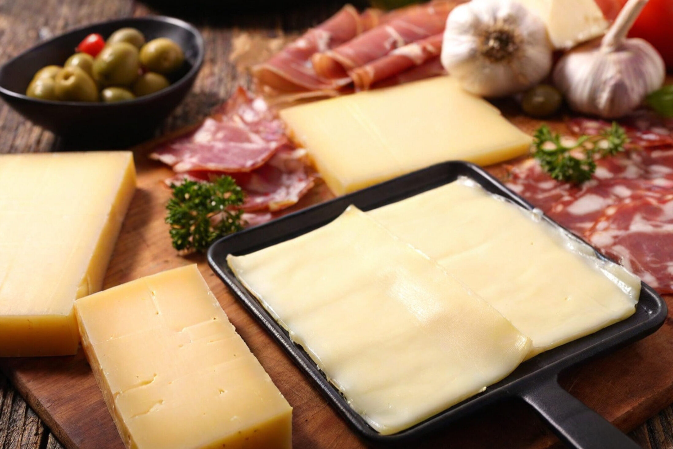 Raclette di formaggi, circondata da pezzi di formaggio, salumi, olive e aglio