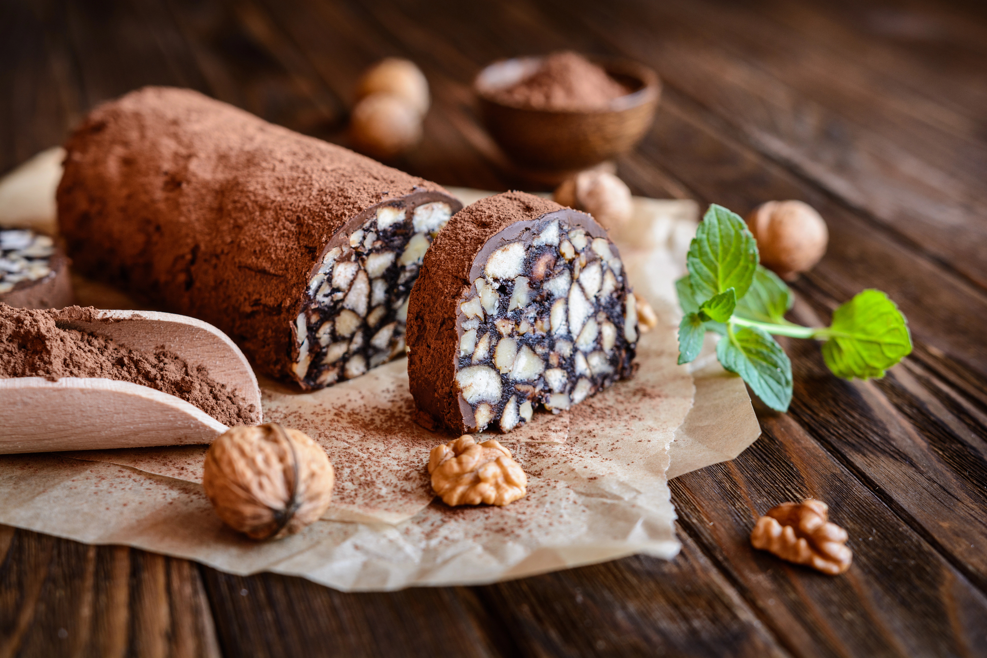 Salame al cioccolato con un fetta tagliata su un pezzo di carta forno su un tavolo con paletta di cacao, frutta secca e rametto di menta; concept: dolci, dessert