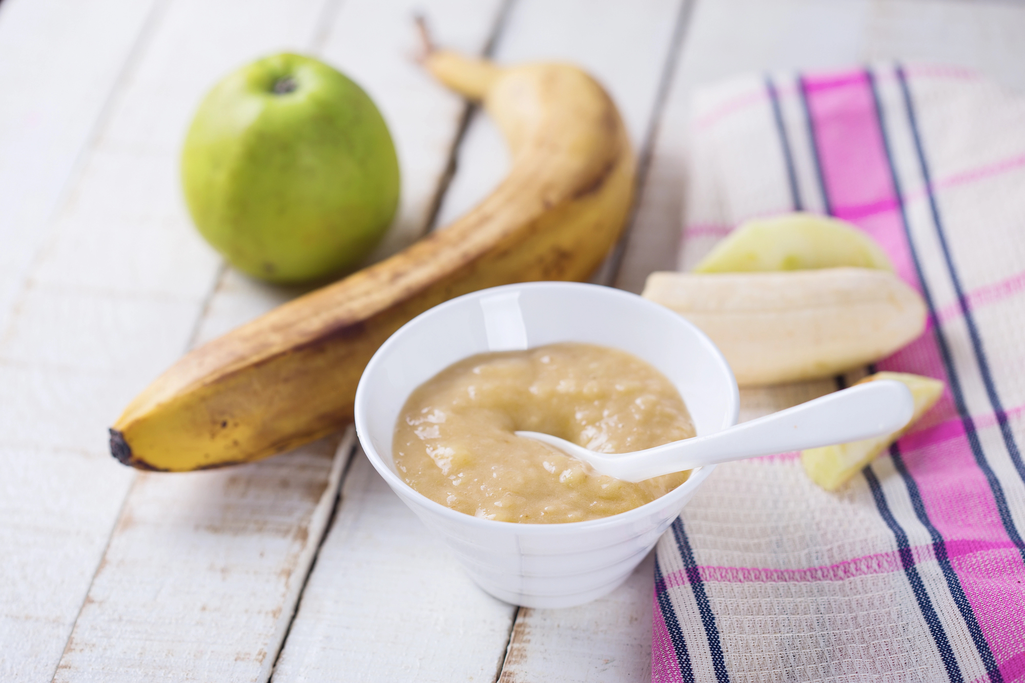 Ciotolina di polpa o purea di mela e banana con cucchiaino, davanti a una mela e a una banana intere e a pezzi di mela e banana; concept: baby food, omogeneizzato