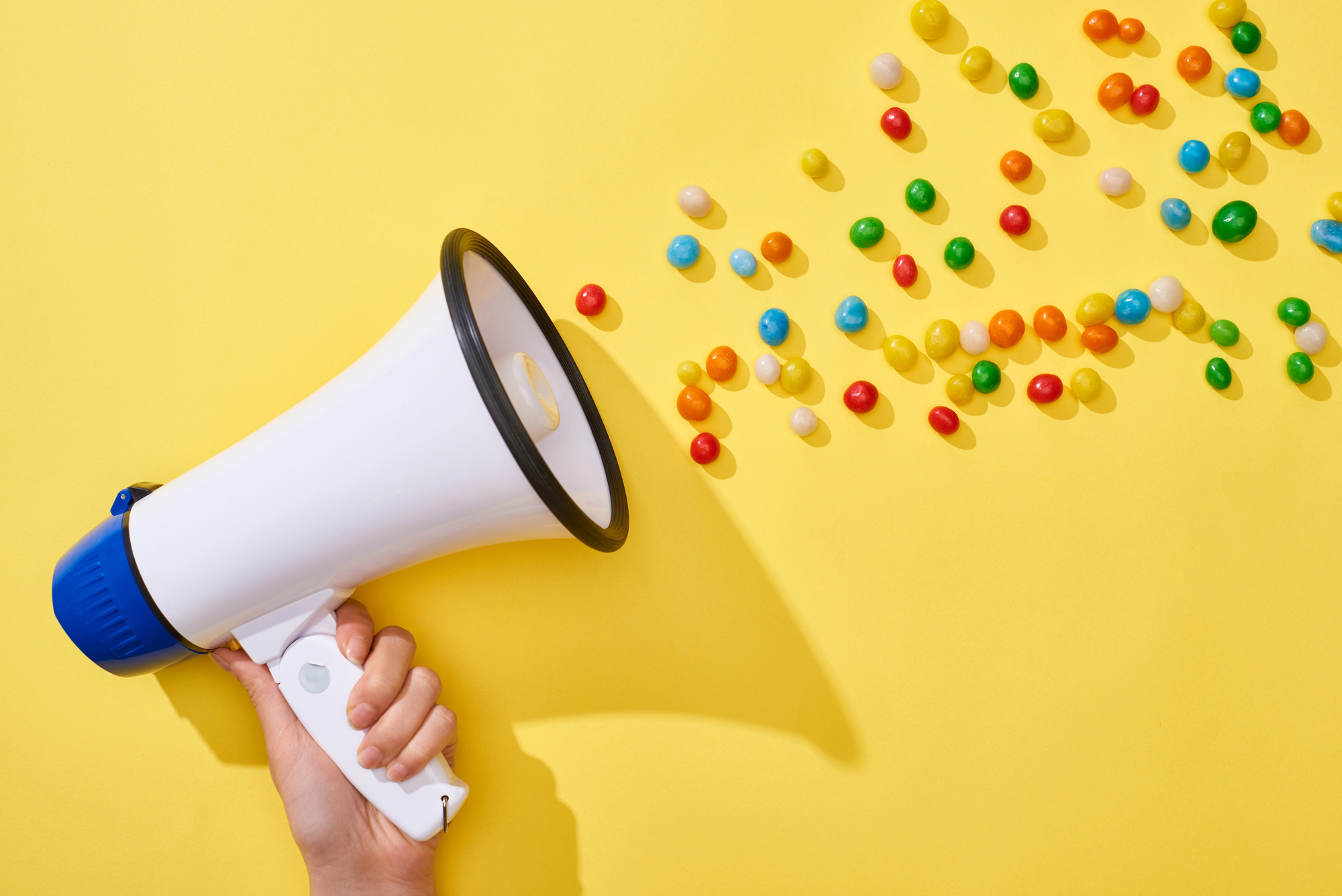 Una mano tiene un megafono su sfondo giallo con caramelle o confetti; concept: allerta alimentare, sicurezza alimentare