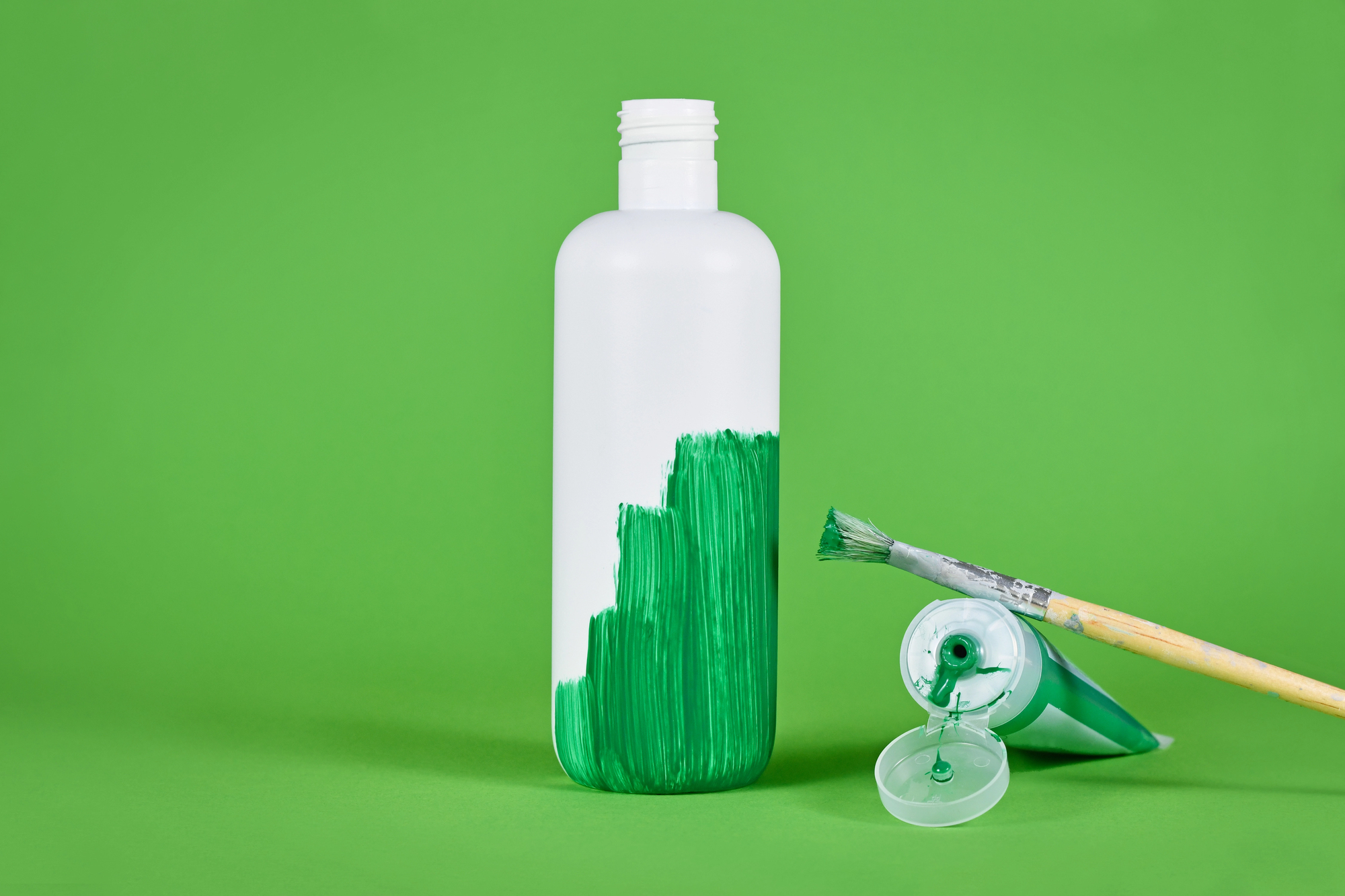 Una bottiglia di plastica pitturata di verde per metà, accanto a un pennello e a un tubetto di tempera verde; conept: greenwashing, green claim