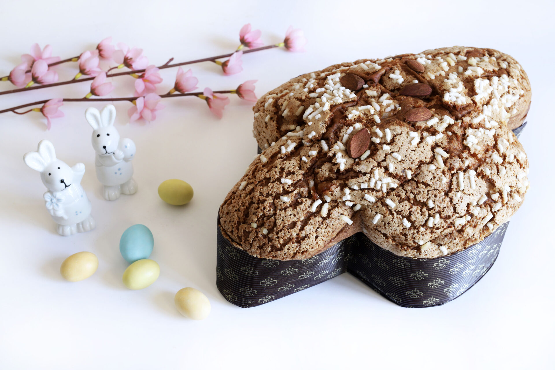 Colomba pasquale glassata e mandorlata con accanto decorazioni pasquali: uova colorate, coniglietti e rami fioriti