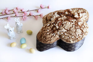 Colomba pasquale glassata e mandorlata con accanto decorazioni pasquali: uova colorate, coniglietti e rami fioriti