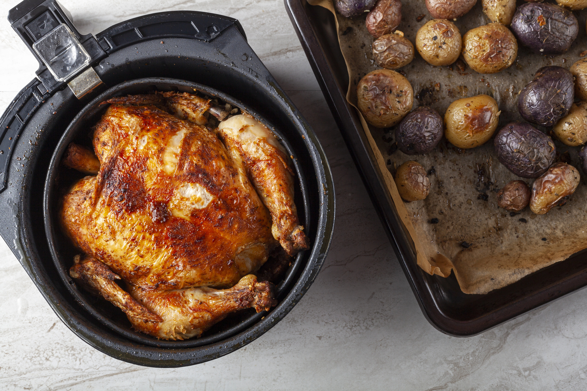Pollo arrostito nel cestello di una friggitrice ad aria, accanto a una teglia di patate al forno su un foglio di carta forno