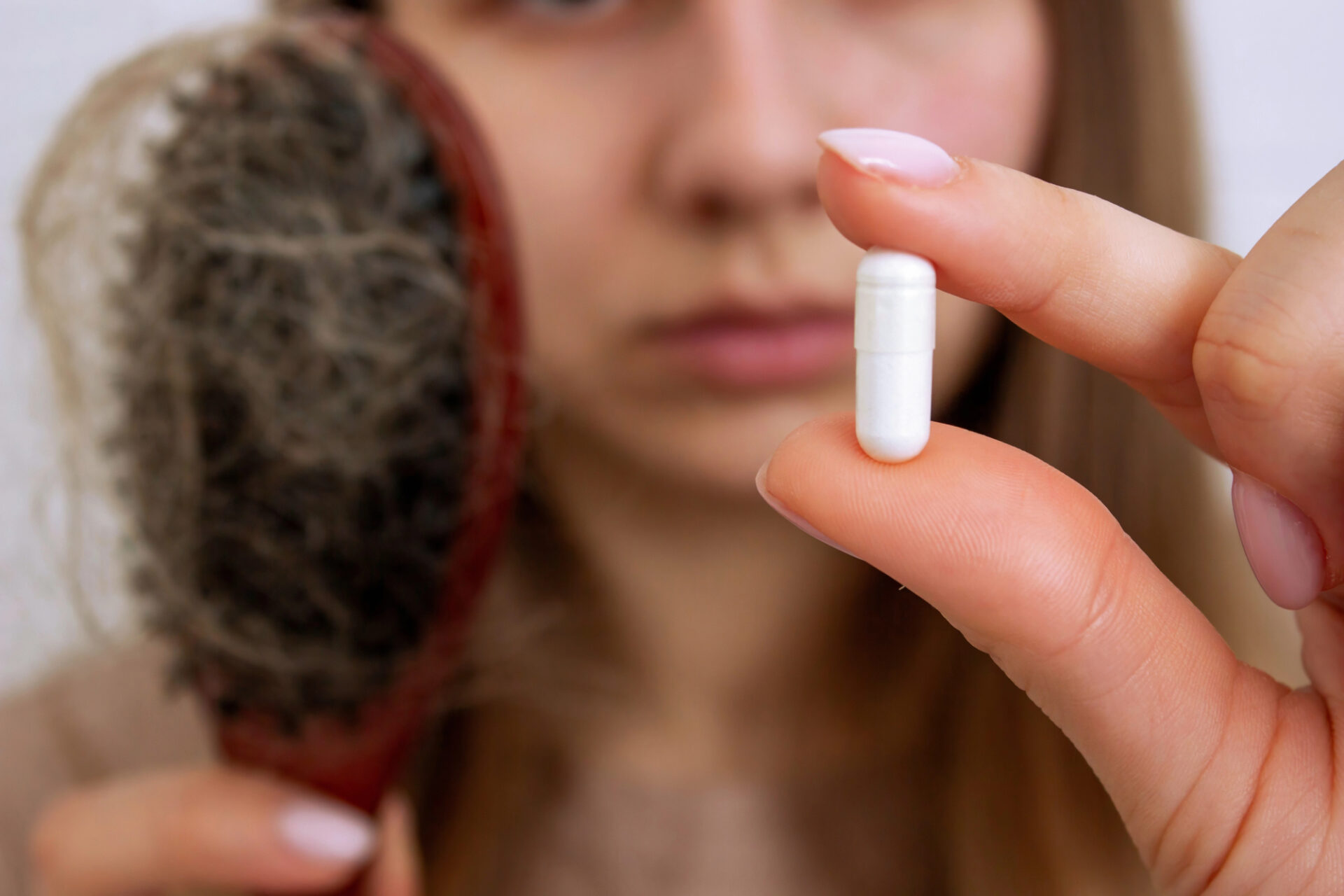 Una donna tiene tra pollice e indice la capsula di un integratore, mentre con l'altra mano tiene una spazzola piena di capelli caduti; concept: integratori alimentari, integratori anticaduta