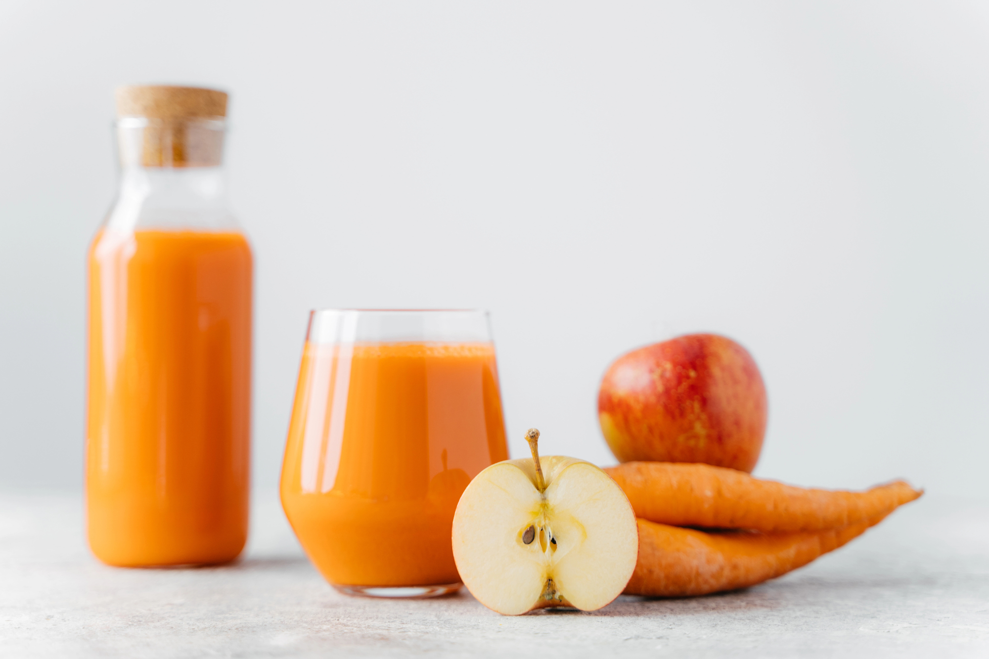 Una bottiglia e un bicchiere di succo di mela e carota, accanto a una mela intera, mezza mela e carote
