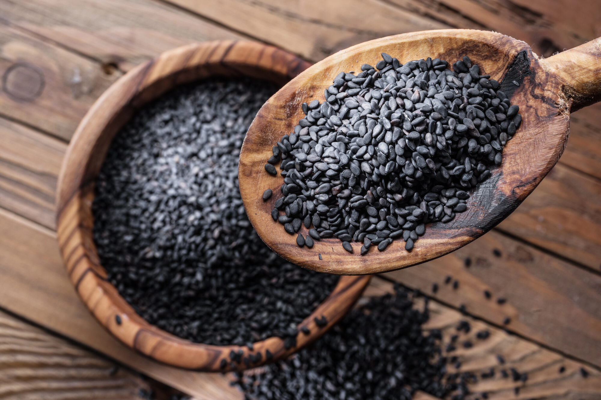 Un cucchiaio di legno pieno di semi di sesamo nero sopra una ciotola di legno piena di semi di sesamo nero