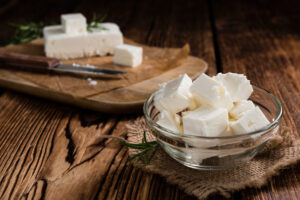 Una ciotola di feta a cubetti, sullo sfondo tagliere con un blocco di feta e un coltello; concept: formaggi dop, latticini