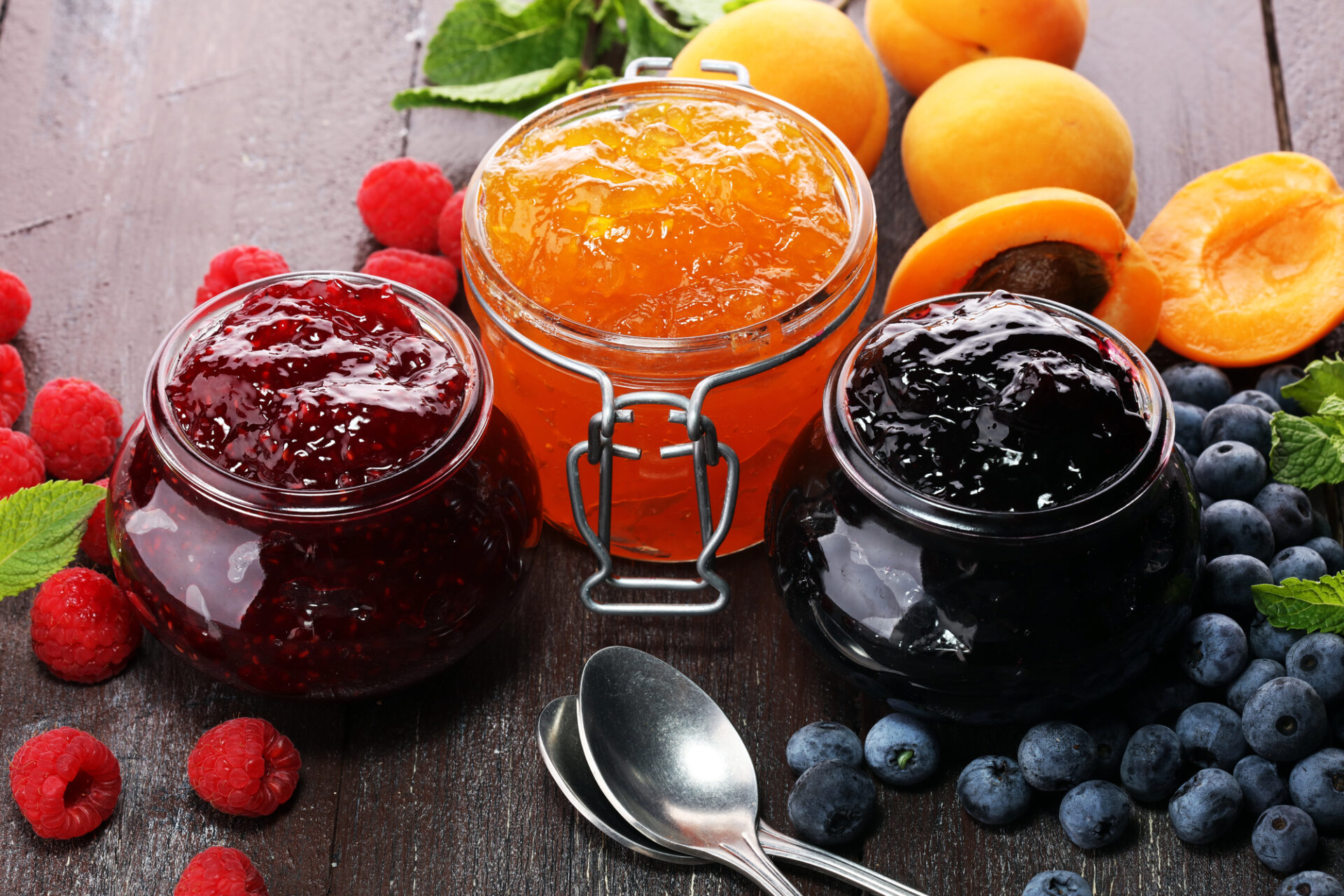 Tre vasetti di marmellate o confetture di lamponi, albicocche e mirtilli, circondate dai frutti freschi