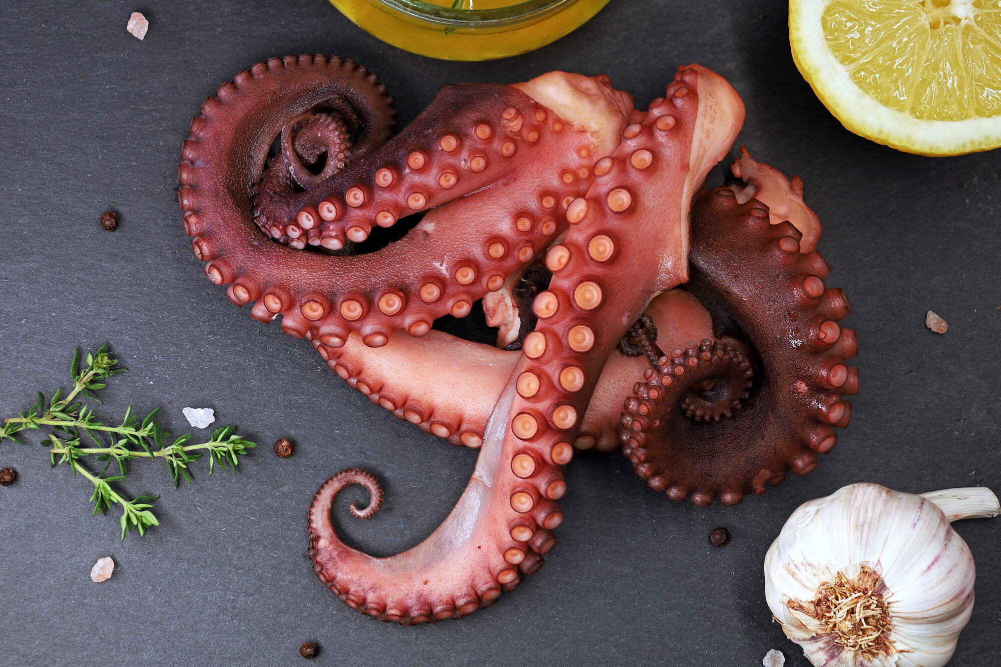 Tentacoli di polpo cotti su ardesia, circondati da erbe aromatiche, sale, grani di pepe, olio di oliva, limone e aglio