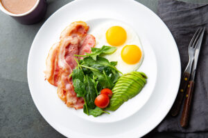 Un piatto con uova all'occhio di bue, fettine di bacon, spinacini, pomodorini e avocado a fette; concept: dieta chetogenica