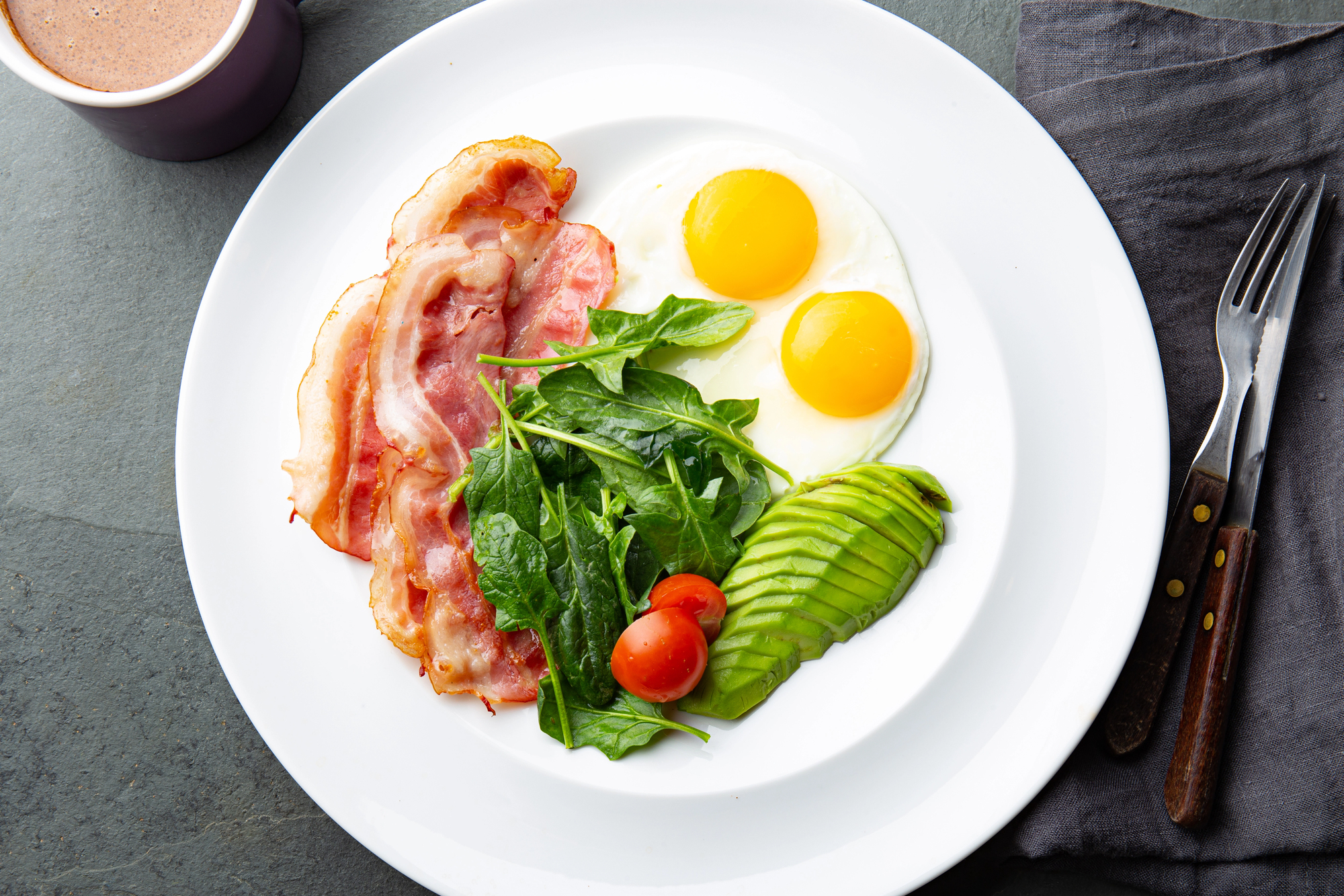 Un piatto con uova all'occhio di bue, fettine di bacon, spinacini, pomodorini e avocado a fette; concept: dieta chetogenica