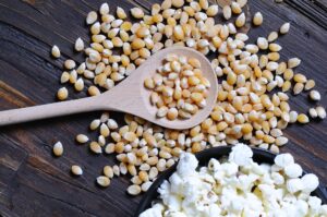 Chicchi di mais per popcorn su un cucchiaio di legno e sparsi su un tavolo, accanto a una ciotola di popcorn