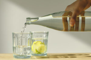 Acqua minerale versata da una bottiglia di vetro in un bicchiere, dietro un altro bicchiere con acqua e fette di limone