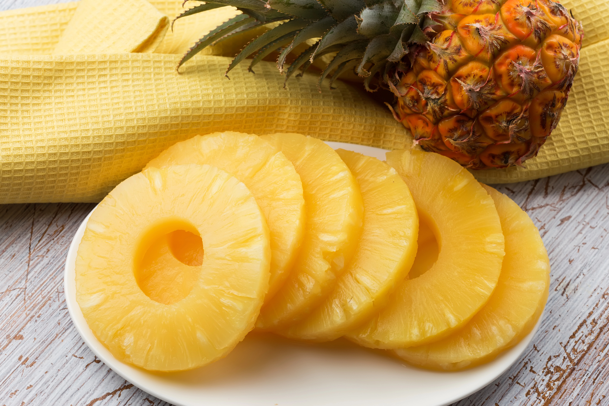 Ananas a fette su un piattino, sullo sfondo un ciuffo d'ananas su un canovaccio giallo